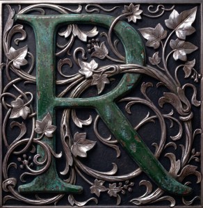 A fancy letter R of weathered metallic green, sits within an entwining silver ivy vine.